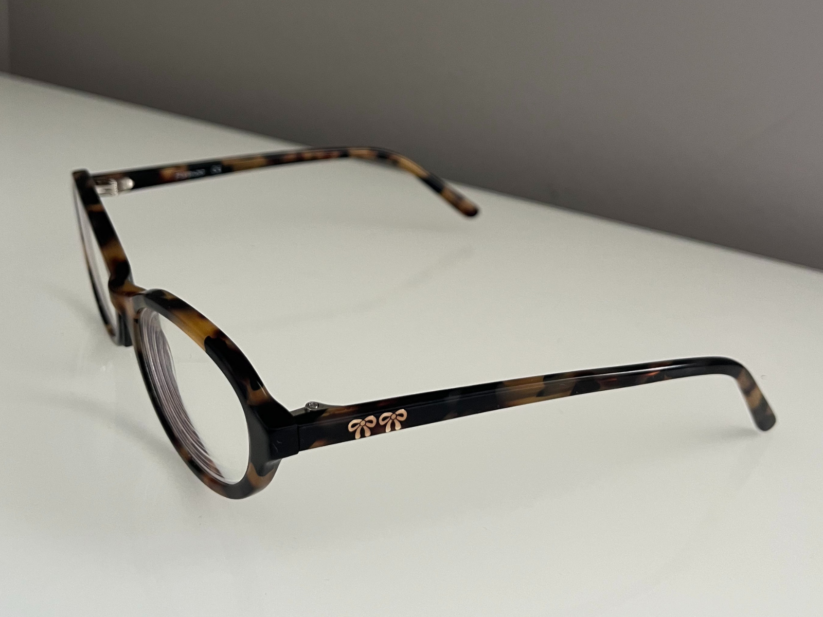 This is another photo of the tortoiseshell glasses, taken from the side angle. The view shows the length and shape of the arms and also the gold details on the temples. The light reflects softly on the shiny surface, showing the brown and black colors of the tortoiseshell pattern and giving depth to the clear lenses.