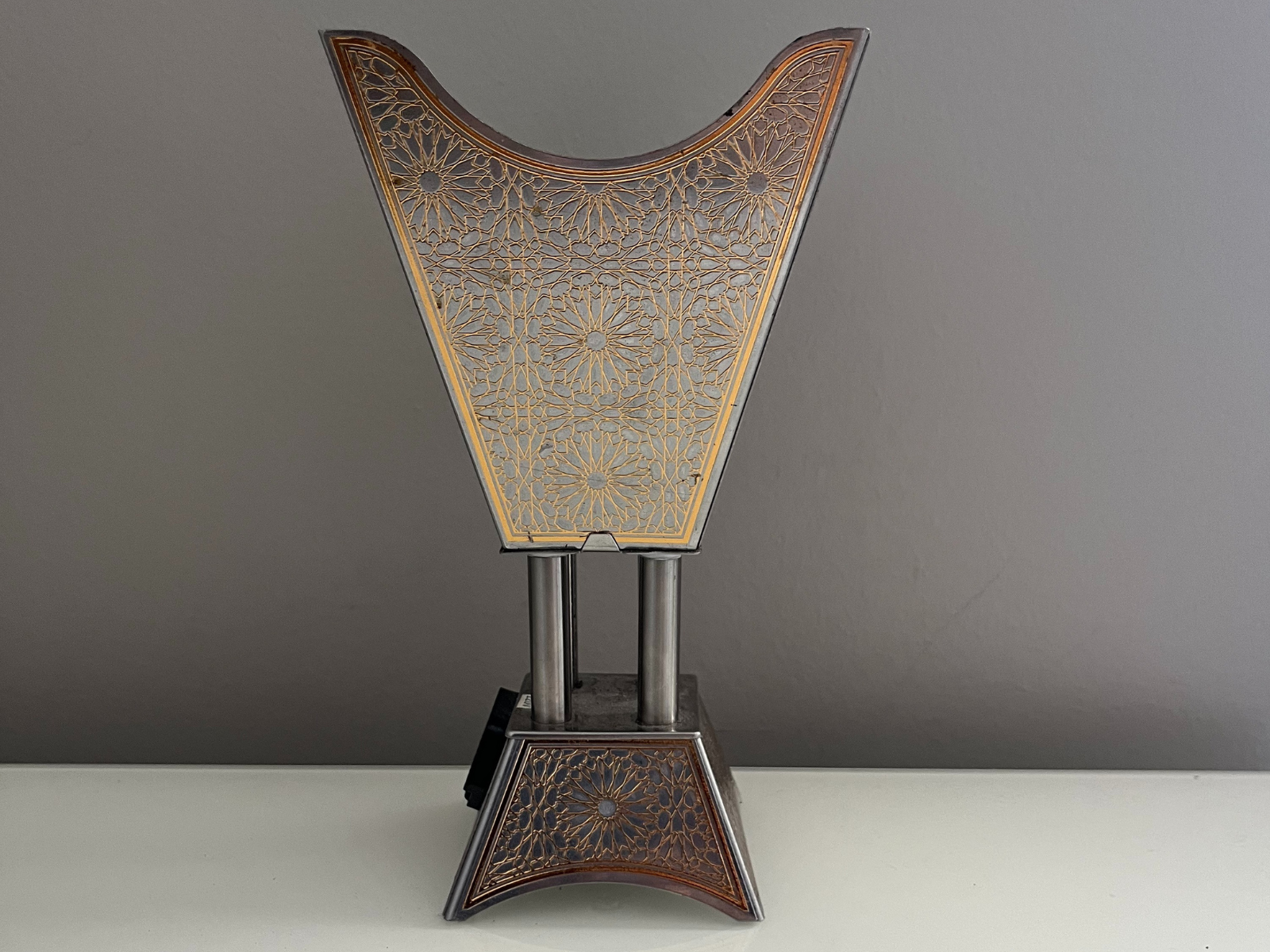 This is a front photo of the bukhoor burner, taken from the center to show its symmetry. The view shows the tall flared top with golden floral patterns, which also repeat on the base. The straight angle makes the geometry clear, from the wide top to the two cylindrical supports and down to the base. The photo shows both the shape and the decoration, making it a good reference for 3D modeling and texturing.