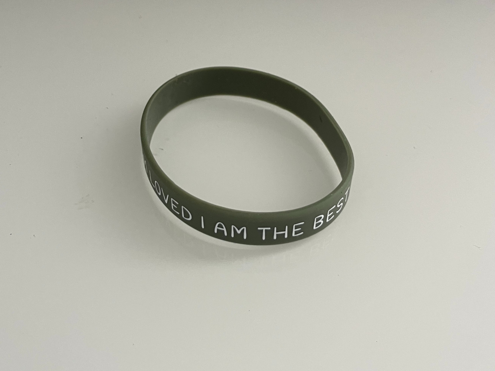 This is a photo of a green silicone wristband on a white surface. The band looks a bit oval, which makes it feel more natural and used, not a perfect circle. The light makes a soft shadow and reflection under the band, which shows its shape. This angle is good for seeing both the curve of the wristband and the printed text clearly.