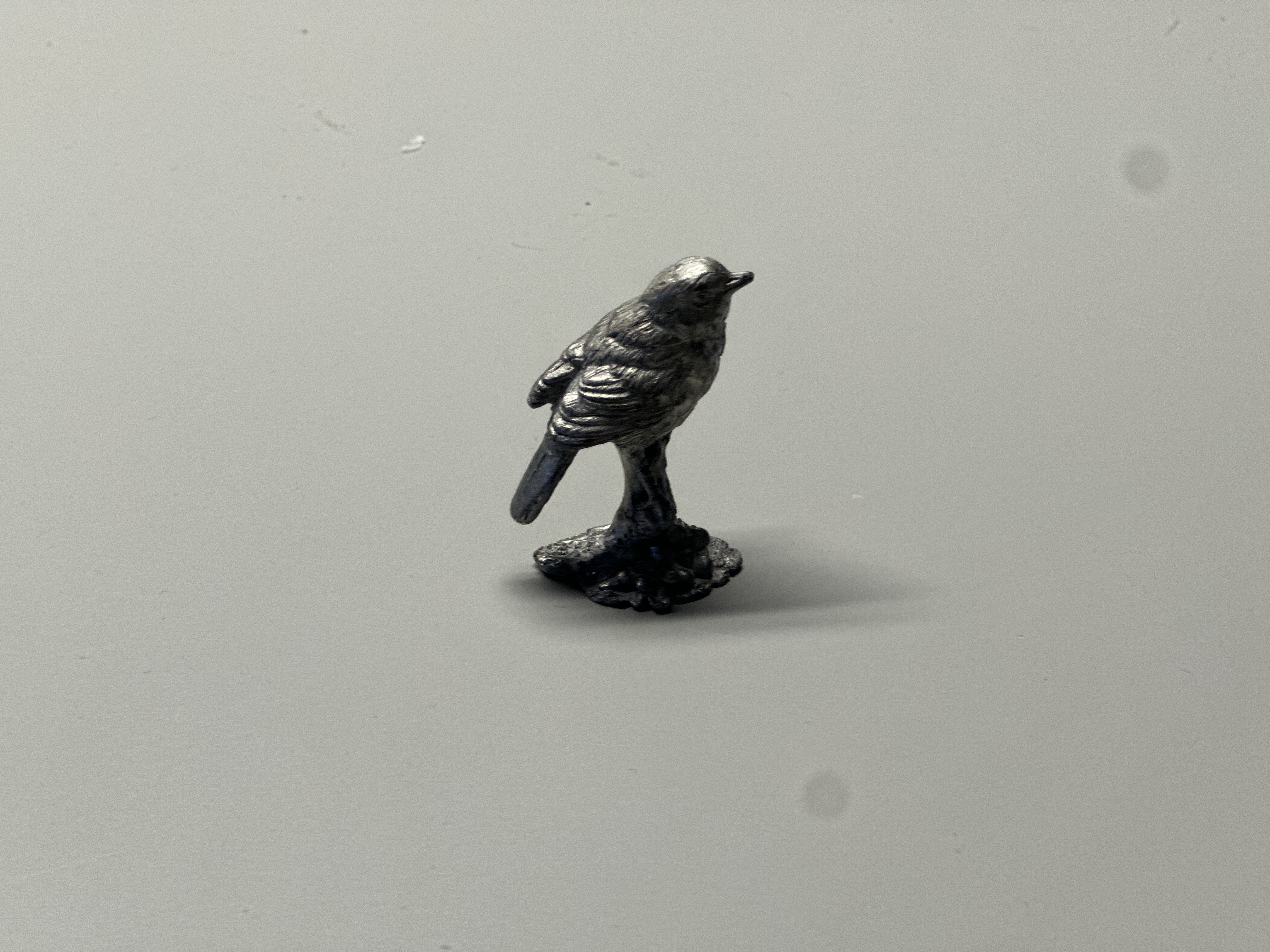 The material is metal, shaped into a small bird sculpture. It is rigid, because metal is strong and does not bend easily. The surface looks refined, with fine details showing the feathers and body texture. It has a metallic shine that makes it look polished and smooth. The dark gray color and solid weight give the piece a durable and elegant appearance.