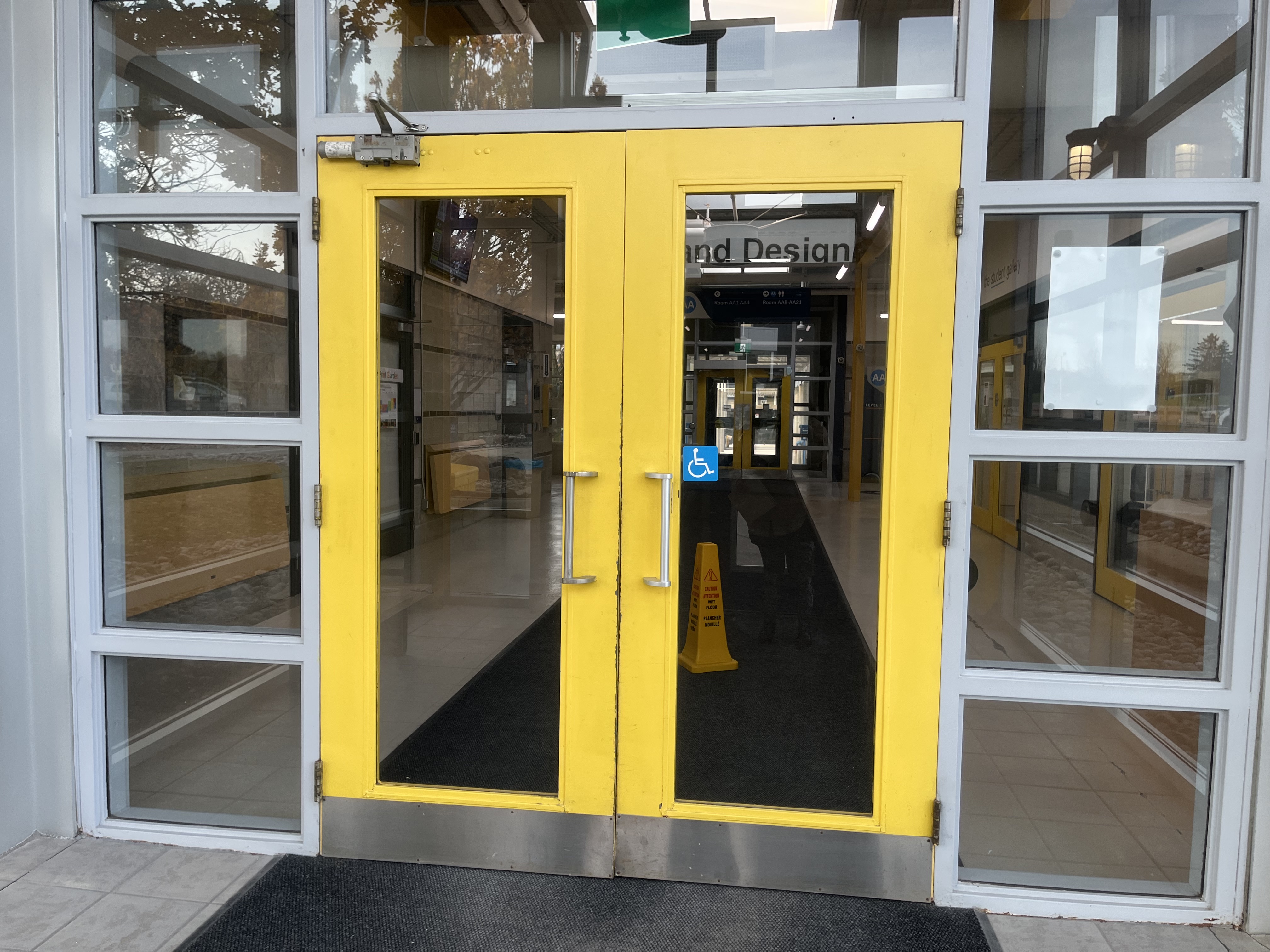 This image shows the main yellow entrance doors leading into the Sheridan Trafalgar Design Wing. The bold yellow color makes the entrance highly visible and welcoming, functioning as both a branding element and a practical navigation cue. The accessibility button is clearly placed, signaling inclusive design considerations for mobility needs. Through the glass, you can see the interior hallway, lockers, signage, and the continuation of yellow trim, reinforcing the visual identity of the building. The entrance acts as a transitional space where students shift from outdoor movement into an academic environment.