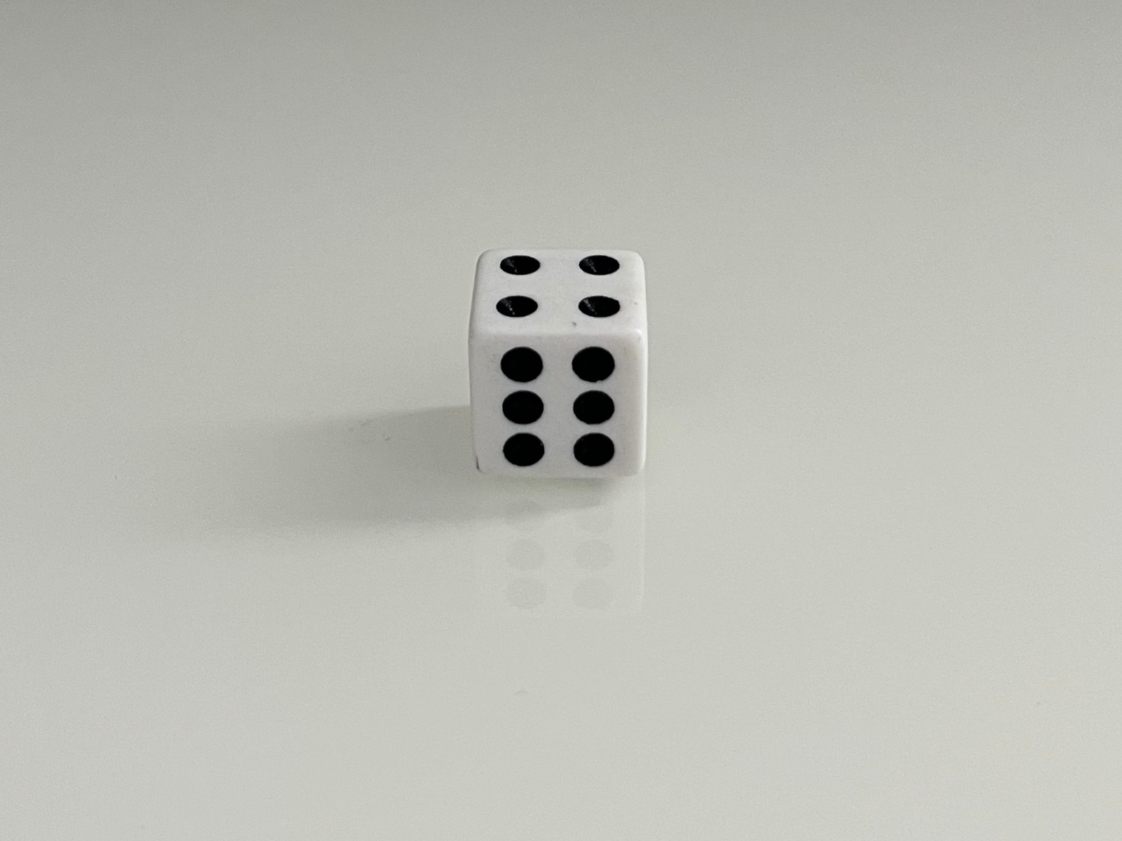 This is a front-facing photo of a white dice, showing the side with six black pips. The angle is more direct and symmetrical compared to the previous image, making the six dots the focal point of the composition. The reflection of the dice on the surface below is clearly visible, reinforcing its three-dimensional form and adding balance to the photo.
