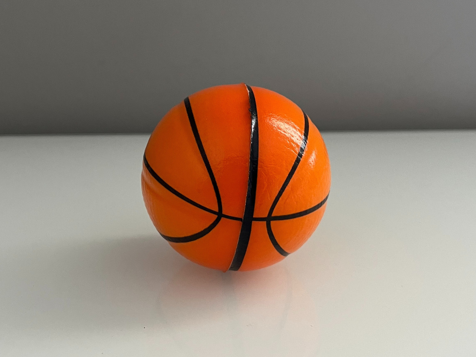 This is a photo of a small basketball, taken at a slight angle that shows the front and side. The shiny surface reflects the light, making the texture look smooth. The black lines curve around the ball and show its round shape. The angle also makes a soft shadow under the ball, which gives the photo more depth.