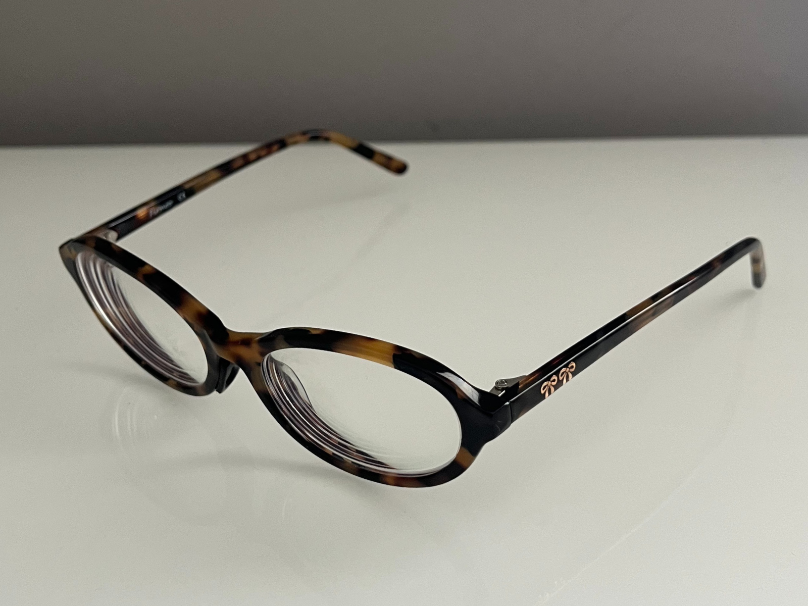 This is a photo of tortoiseshell glasses, taken at a three-quarter angle that shows both the front frame and the side arms. The view highlights the oval lenses and also shows small details in the frame, like the gold parts on the temples. The shiny tortoiseshell texture reflects the light, giving the frame more depth, and the clear lenses also show soft reflections.