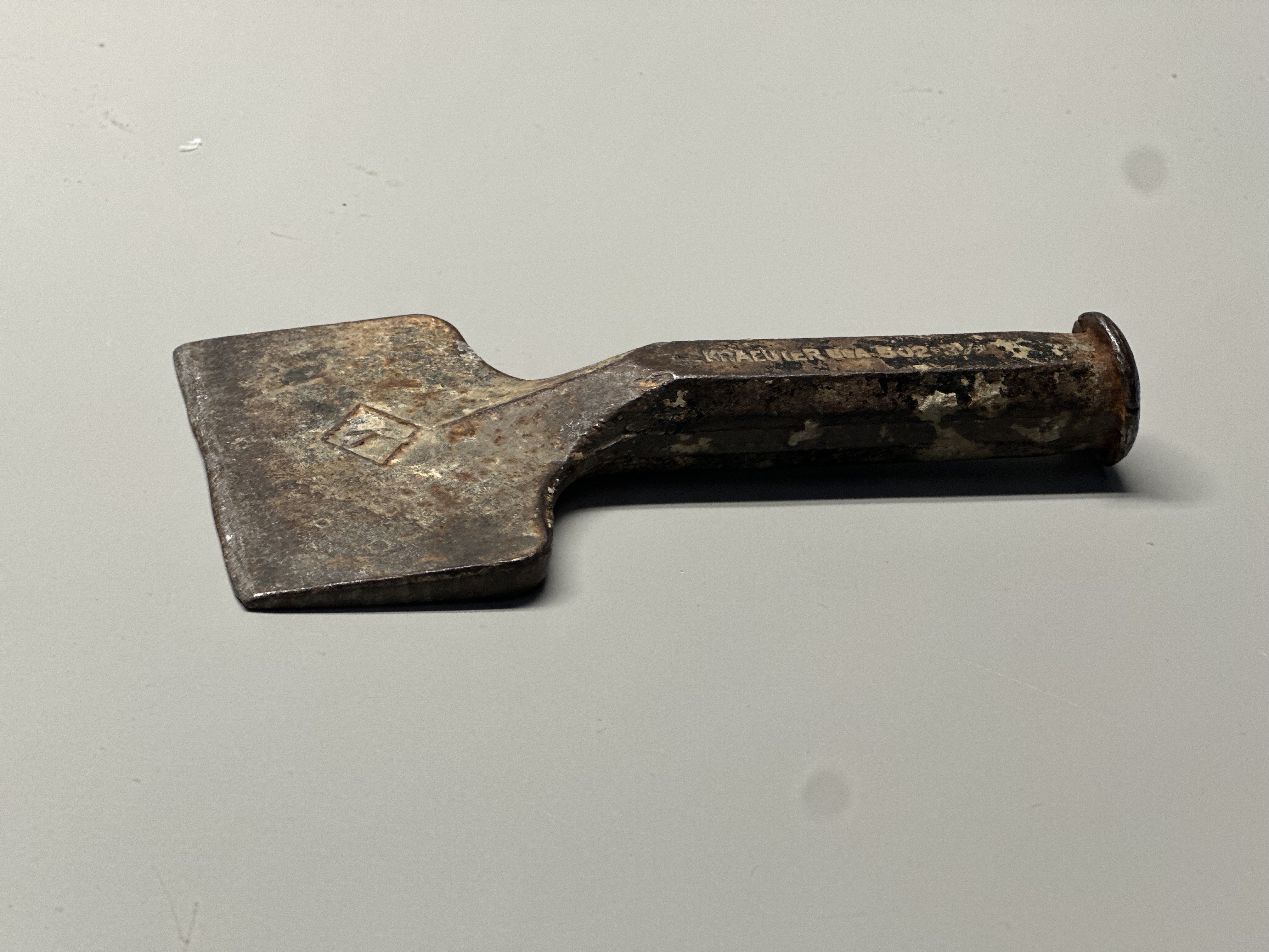 The material is metal, most likely steel or iron, that has been forged and shaped into a tool. It is rigid, because metal is strong and does not bend easily. The surface looks unrefined, with scratches, rust, and marks that show age and use. The texture is rough and worn, giving it an industrial look. The dark color and visible corrosion suggest that it has been exposed to air and moisture over time.